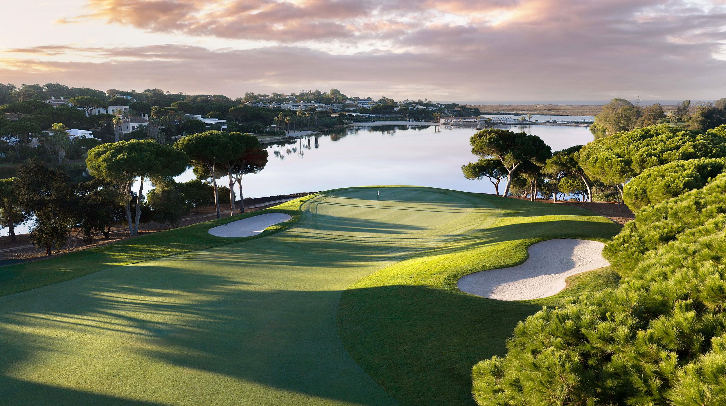 A green framed by umbrella pines and two bunkers beside a lake at sunrise